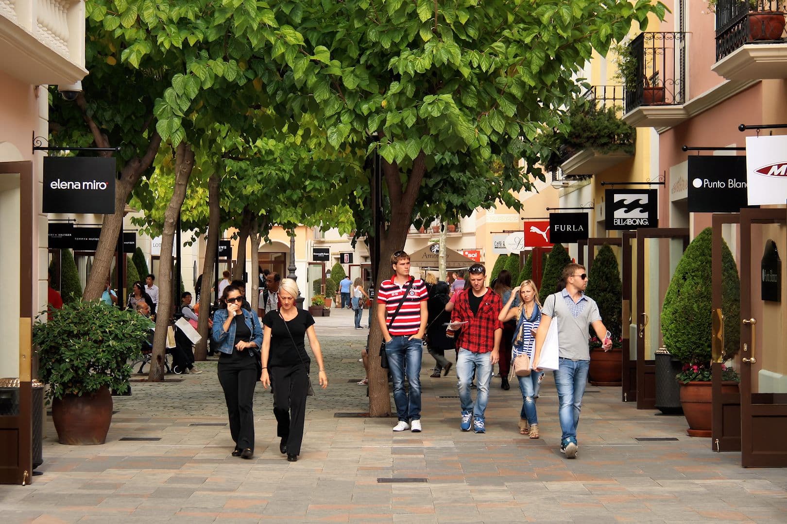 Allée piétonne arborée de La Roca Village, bordée de boutiques de marques avec leurs enseignes noires