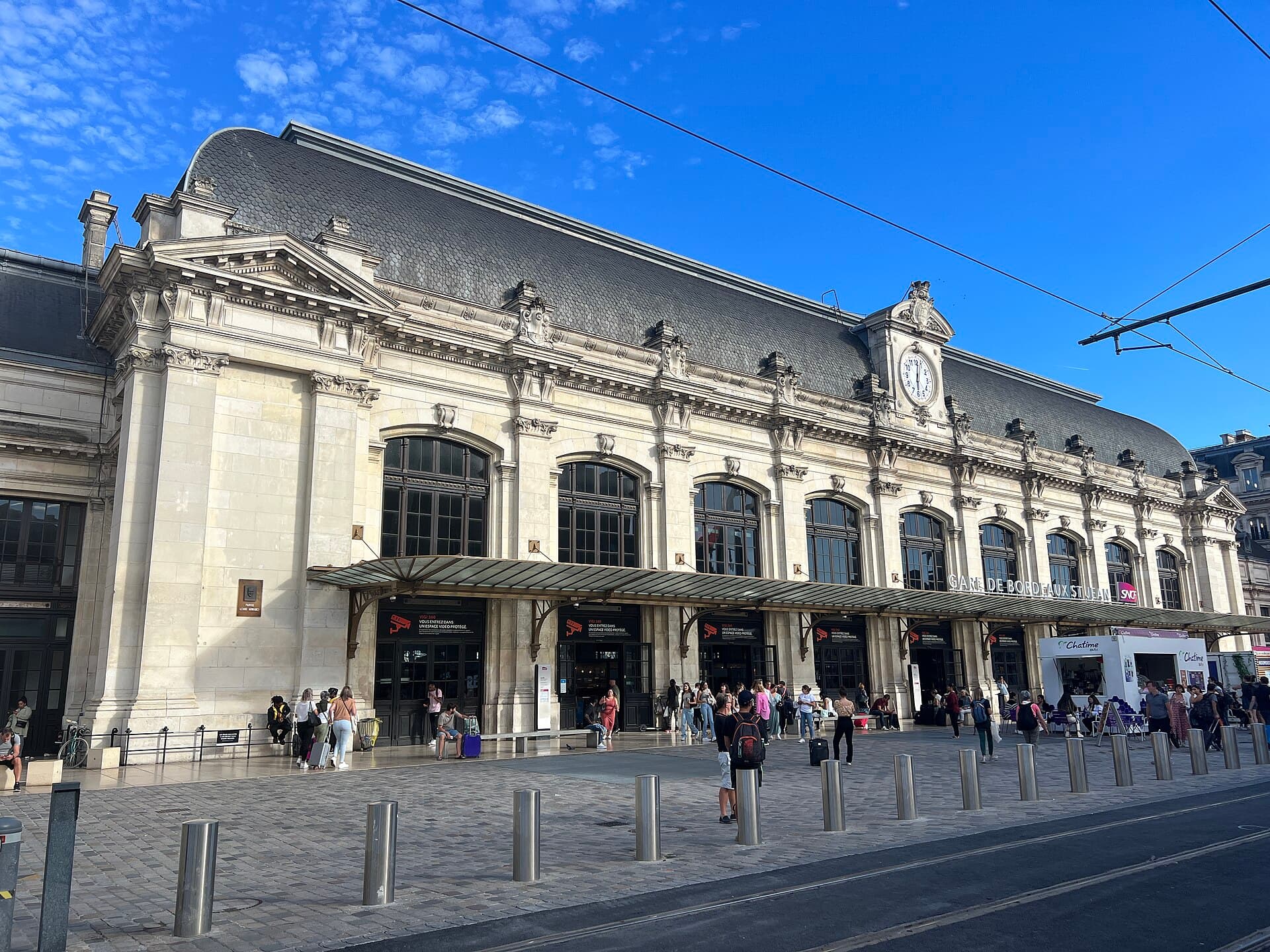 Façade en pierre et horloge de la gare de Bordeaux-Saint-Jean, voyageurs avec valises sur le parvis